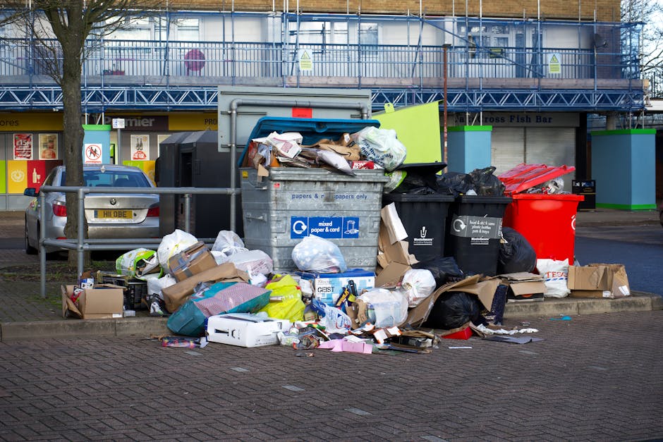 In the foreground, a large pile of mixed rubbish, including black and black plastic waste bags, flattened cardboard boxes, crumpled paper, and various packaging materials, is gathered on a paved sidewalk next to a metal railing. Some waste bags are torn open, revealing their contents, while other items such as cardboard boxes and paper are scattered around the pile. Behind this, there is a grey, industrial-style wheelie bin labeled for mixed paper and cardboard, which is overflowing with additional bags and loose waste items stacked on top and spilling over the sides. To the left of the bin, a small silver car with a visible license plate is parked, partially obstructed by the rubbish. In the background, a row of shops with brightly colored signage and a shopfront with a yellow awning can be seen, along with a building covered in scaffolding and protective blue netting, indicating ongoing construction or maintenance work. The scene is captured during daylight under overcast skies, highlighting the contrast between the urban environment and the unmanaged waste collection scene, typical of private rubbish collection services provided by local waste management companies like Rubbish Removal Streatham.