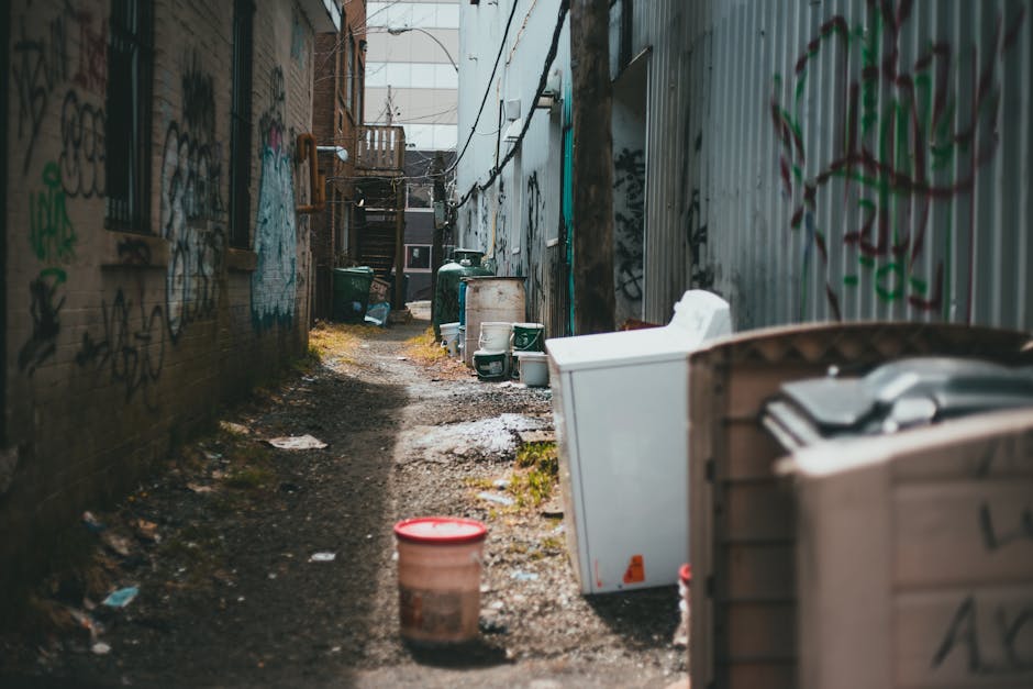 The image depicts a narrow alleyway with uneven, muddy ground, scattered with patches of water and debris. On the right side, there are large outdoor appliances including an upright washing machine and a refrigerator, both appearing used and slightly weathered, positioned close to a corrugated metal fence covered in graffiti. On the left, a brick wall with visible mortar joints is also marked with graffiti, and small, barred windows are set into the wall. Towards the background, several green wheelie bins are lined up near the base of wooden stairs leading to an upper-level entrance of a building, and additional waste containers are visible further along the alley. The environment is lit naturally, suggesting daylight, and the overall scene presents an urban setting where private waste disposal objects are temporarily stored, aligning with typical scenes requiring professional rubbish removal services such as those offered by Rubbish Removal Streatham, especially in areas with restricted access for standard waste collection.