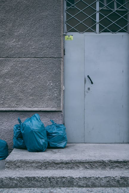 The image shows four blue plastic rubbish bags placed on a concrete step in front of a grey metal door with a black handle, situated at the base of a building with a textured, grey exterior wall. To the right of the bags, a small yellow warning sticker is visible on the wall near the door. The door features a diamond-patterned window grille above it, with glass panes allowing light to pass through. The scene appears to be outdoors, on a city pavement or alleyway, with the bags containing waste awaiting collection or disposal. The environment suggests an example of private or independent waste handling, commonly associated with rubbish removal services. The overall atmosphere is neutral and functional, emphasizing the practical aspect of rubbish collection in an urban setting, as performed by companies like Rubbish Removal Streatham, who handle various waste clearance tasks efficiently and professionally.